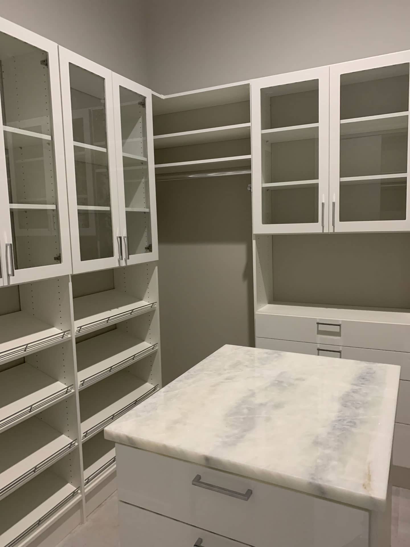 White walk-in closet with glass-front upper cabinets, angled shoe shelves and a center island with a marble-look top and drawers