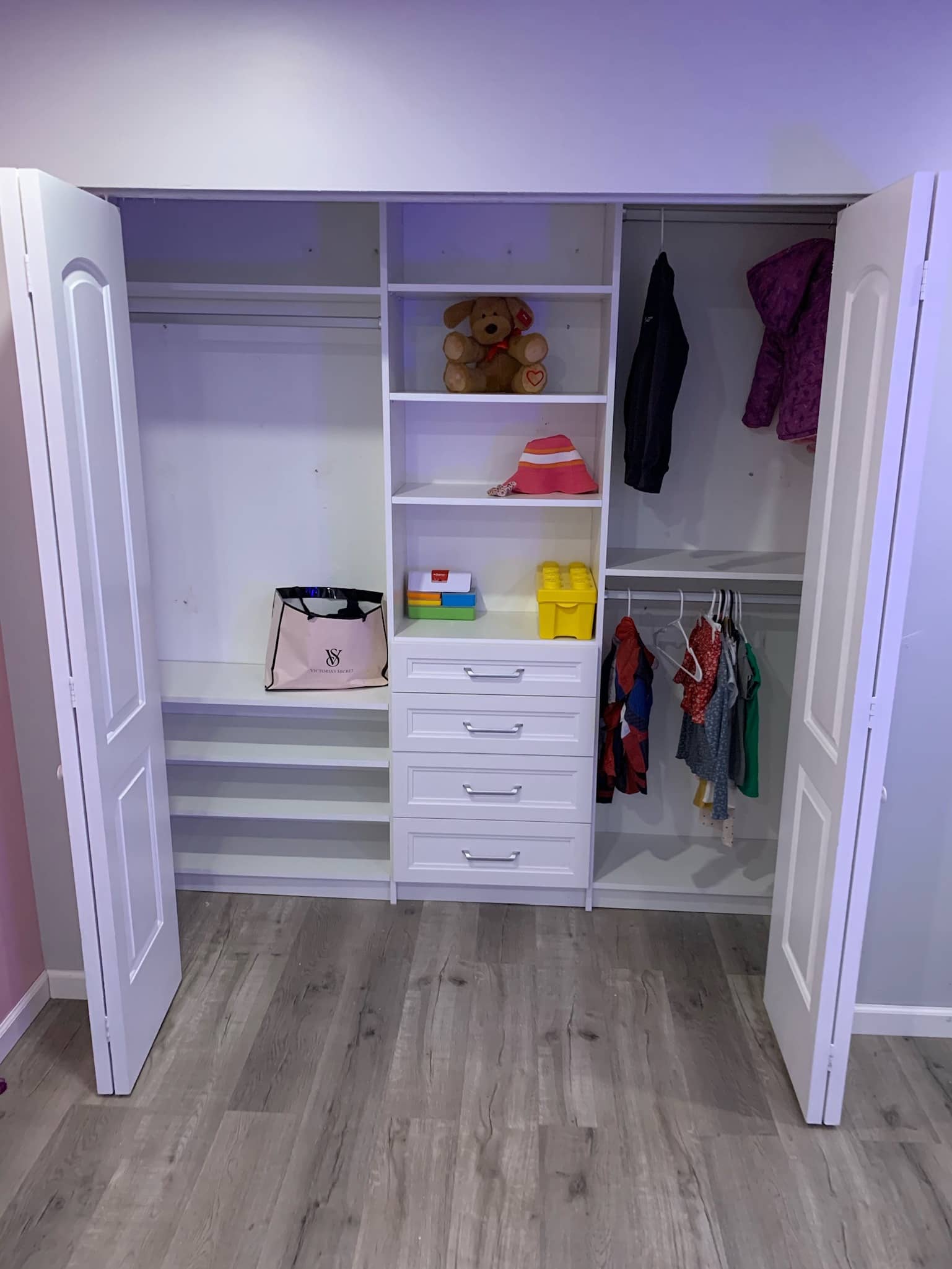 Child's reach-in closet in white with a center tower of shelves and drawers, a low rod of kids' clothes and a teddy bear on a shelf