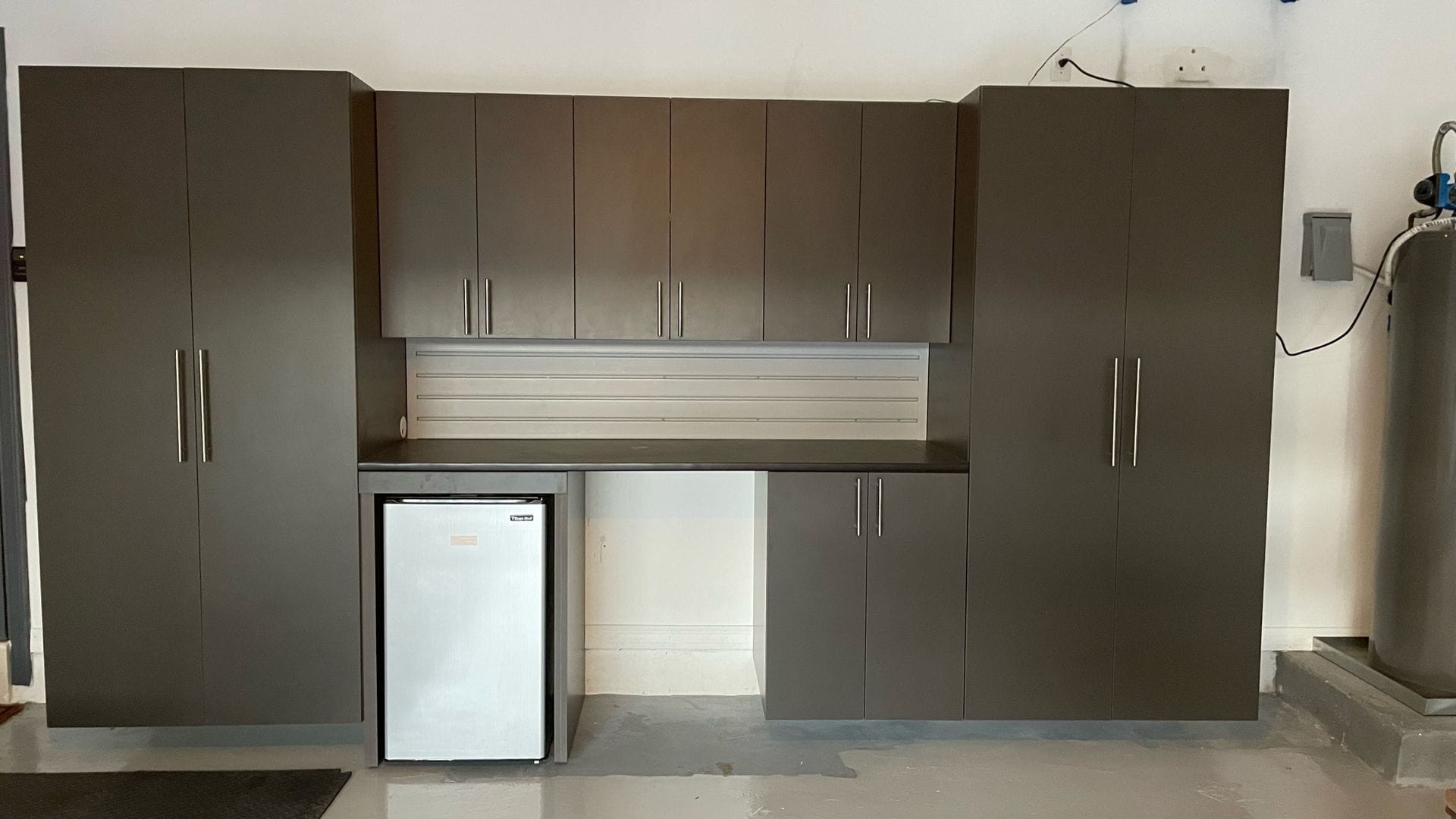 Garage wall of dark gray cabinets with tall storage units, a workbench with a slatwall backsplash and an open bay for a mini fridge