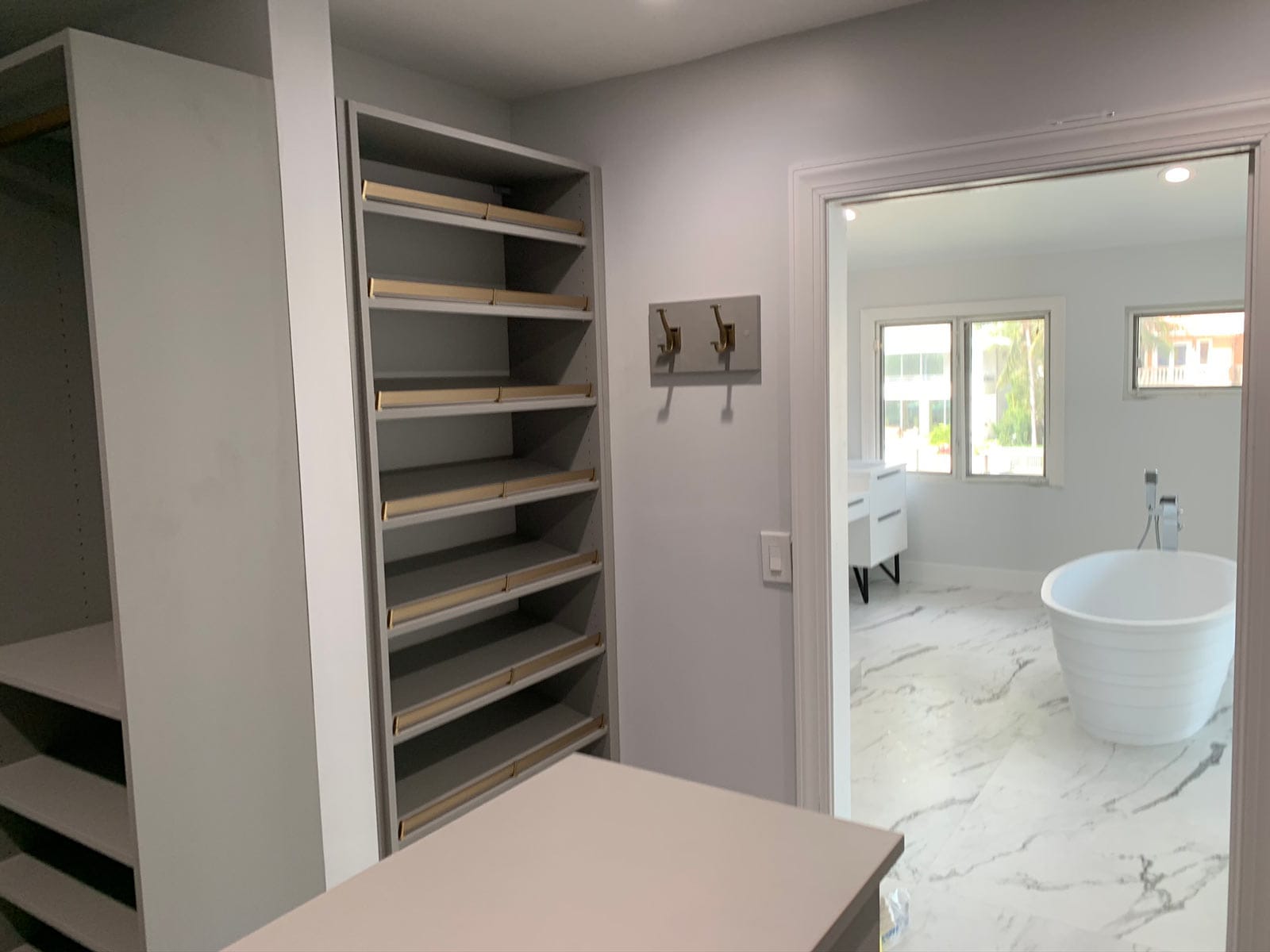 Gray closet with slanted shoe shelves trimmed in gold, a wall hook rack and an island, opening onto a marble bathroom