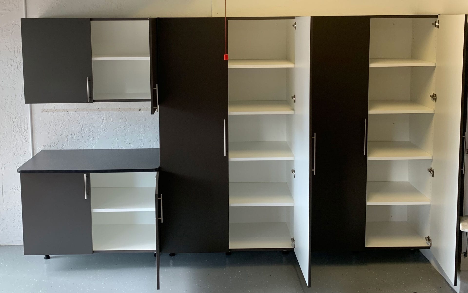 Open garage cabinets showing adjustable white shelves inside charcoal cabinet boxes, with a countertop section on the left