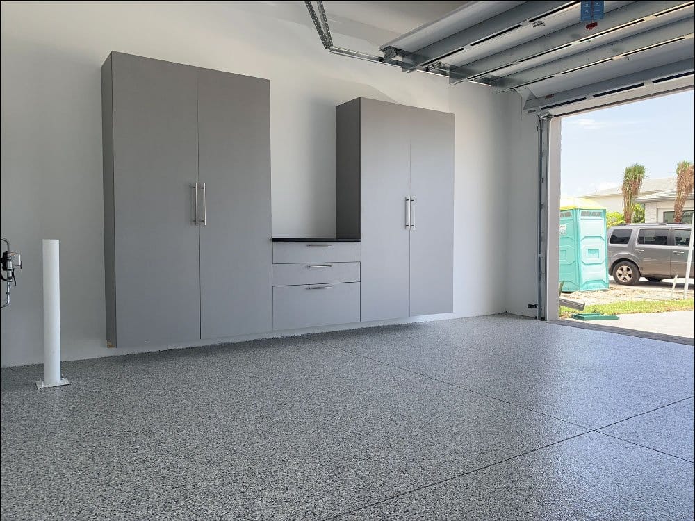 Gray garage cabinets with a drawer bank on a gray flake epoxy floor, with the garage door open to a sunny street