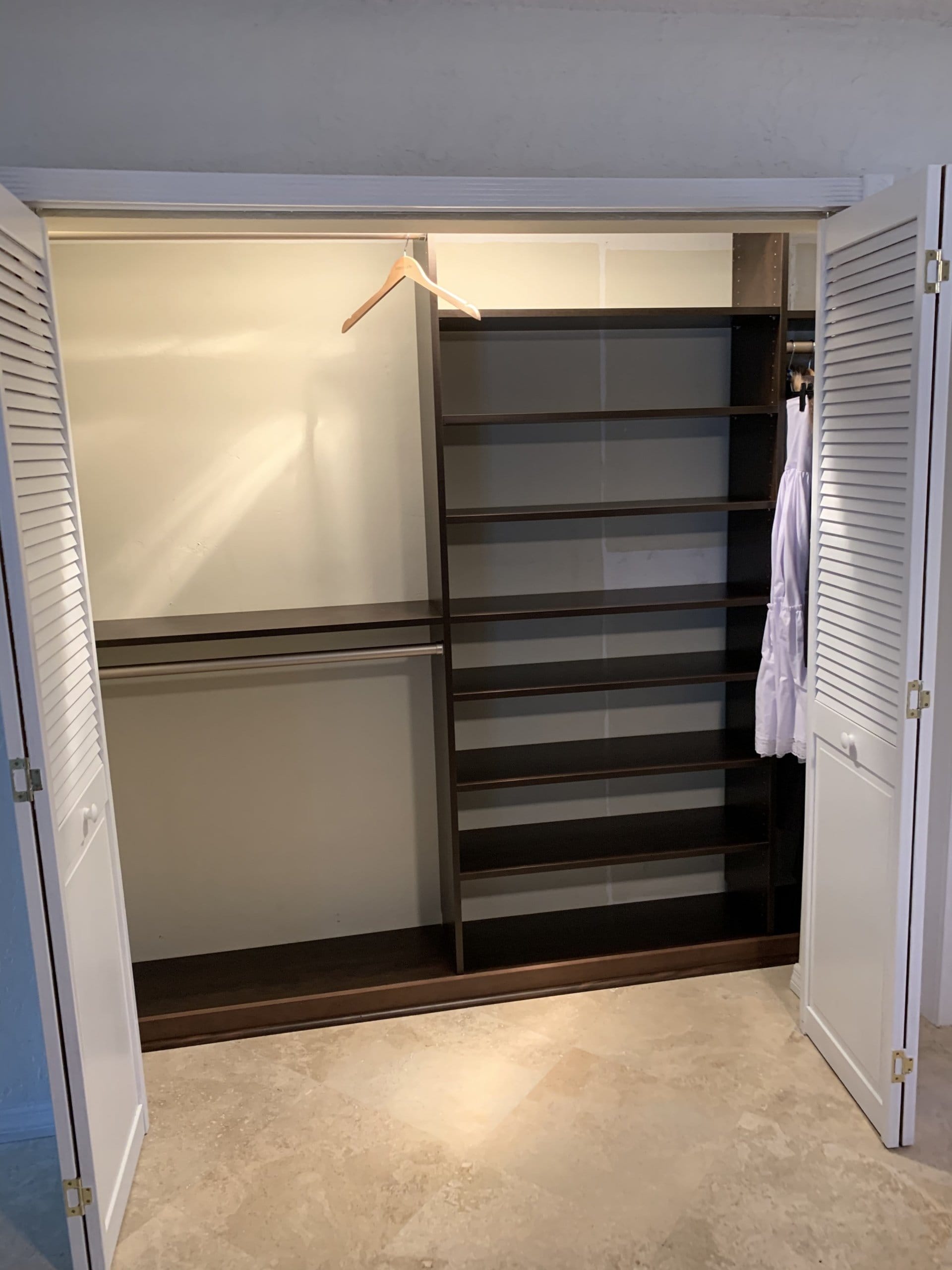 Dark wood-grain reach-in closet behind louvered bifold doors, with double-hang rods on the left and a tall adjustable shelf tower on the right