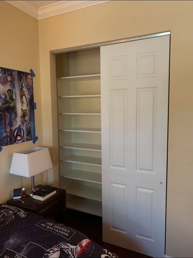 Kid's bedroom closet with full-height adjustable white shelves behind a sliding door, next to a superhero poster on the wall