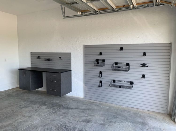 Gray workbench with a slatwall backsplash beside a slatwall panel holding wire baskets and hooks in a garage